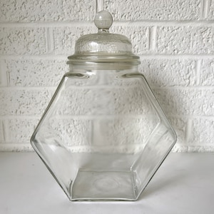 May include: A clear glass jar with a hexagonal shape and a glass lid. The jar has a smooth, rounded top and a knob on the lid.
