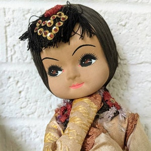 Op de afbeelding: Een vintage pop met een geschilderd gezicht, donker haar en een decoratieve hoofdtooi versierd met gouden pailletten. De pop draagt een tan en goudkleurige jurk met een rood en goud geborduurde kraag. De pop is geplaatst tegen een witte bakstenen muur.