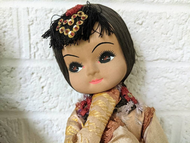 Op de afbeelding: Een vintage pop met een geschilderd gezicht, donker haar en een decoratieve hoofdtooi versierd met gouden pailletten. De pop draagt een tan en goudkleurige jurk met een rood en goud geborduurde kraag. De pop is geplaatst tegen een witte bakstenen muur.