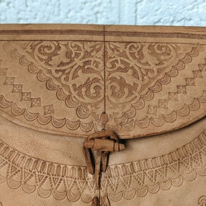 Puede incluir: Un bolso de cuero marr&oacute;n claro con intrincados dise&ntilde;os en relieve. El bolso tiene un cierre de solapa con una correa de cuero decorativa y un borla.