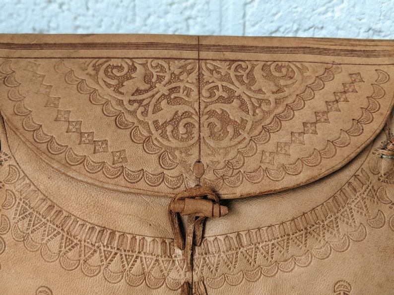 Puede incluir: Un bolso de cuero marr&oacute;n claro con intrincados dise&ntilde;os en relieve. El bolso tiene un cierre de solapa con una correa de cuero decorativa y un borla.