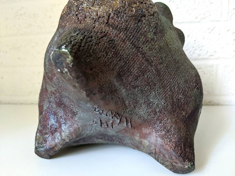 May include: A dark brown, textured ceramic sculpture with a rough, uneven surface. The sculpture is signed "Becky Smith" on the bottom.