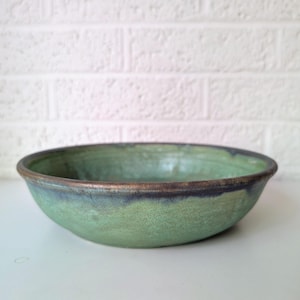 May include: A handmade ceramic bowl with a wide, shallow design. The bowl features a green glaze with a dark blue rim and a bronze-colored edge. The bowl is set against a white background and a white brick wall.