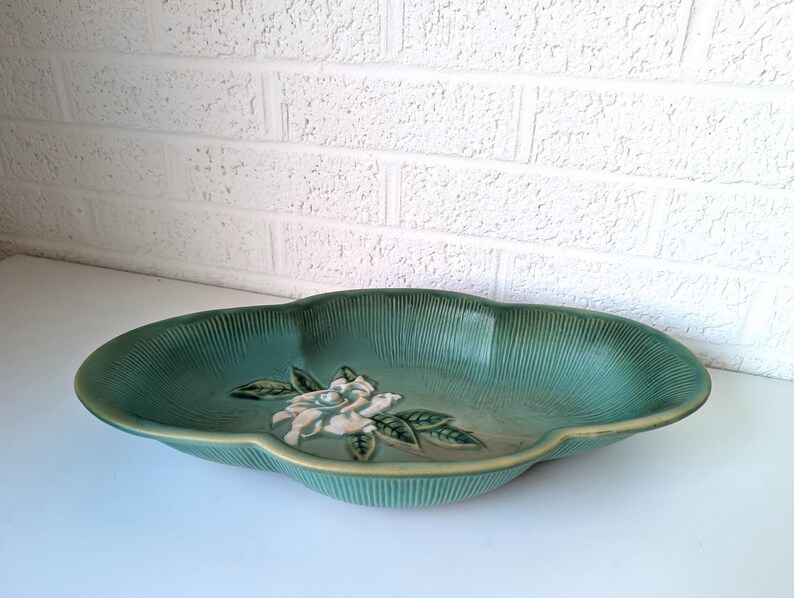 Puede incluir: Un plato de servir de cer&aacute;mica verde con un dise&ntilde;o de flor blanca en relieve. El plato tiene un borde festoneado y es perfecto para servir aperitivos o aperitivos.
