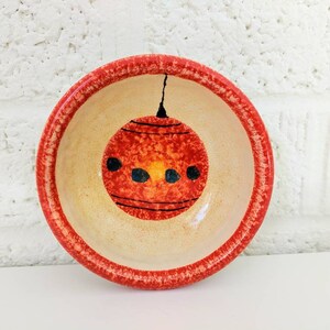 May include: A small, red and white ceramic bowl with a hand-painted design of a red Christmas ornament. The bowl has a red rim and a white interior.