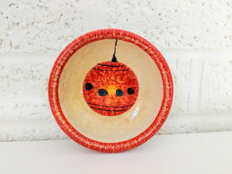 May include: A small, red and white ceramic bowl with a hand-painted design of a red Christmas ornament. The bowl has a red rim and a white interior.