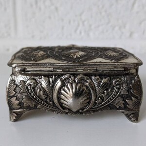 May include: A silver metal trinket box with an ornate design. The box has a hinged lid and sits on four feet.