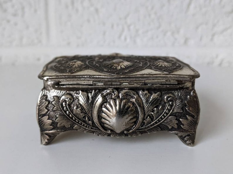 May include: A silver metal trinket box with an ornate design. The box has a hinged lid and sits on four feet.