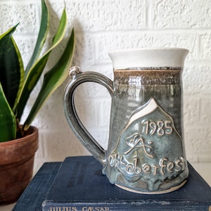 May include: A green and brown ceramic beer stein with a white rim and handle. The stein has a mountain scene with the text "1985 Oktoberfest" on it.