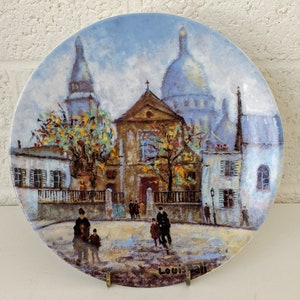 Op de afbeelding: Een vintage keramische plaat met een geschilderd tafereel van een Parijse straat met gebouwen, bomen en mensen. De plaat is wit met een blauw en bruin geschilderd ontwerp. De tekst "LOUIS XII" is zichtbaar aan de onderkant van de plaat.