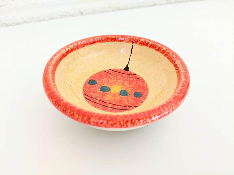 May include: A small, round ceramic dish with a red rim and a hand-painted design of a red Christmas ornament with blue and yellow accents.