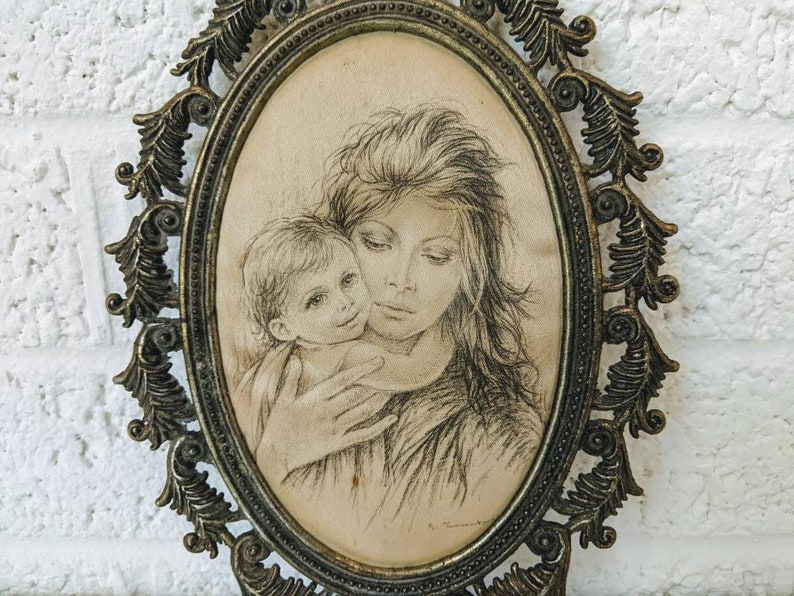 May include: A vintage oval-shaped framed drawing of a mother and child. The drawing is in black and white and depicts a woman holding a young child. The frame is ornate and has a gold finish.