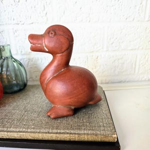 Pode incluir: Uma figura de pato de madeira esculpida à mão com um acabamento marrom avermelhado. O pato está sentado em uma superfície texturizada.