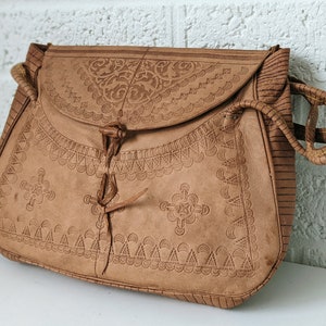 Puede incluir: Un bolso de cuero marr&oacute;n con intrincados dise&ntilde;os florales y geom&eacute;tricos en relieve. El bolso tiene un cierre de solapa con un cordel de cuero y una correa de cuero trenzada.