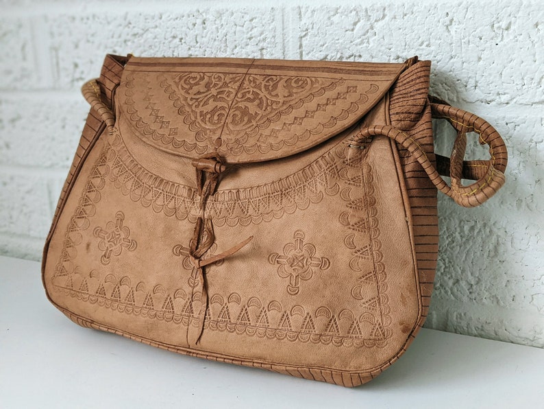 Puede incluir: Un bolso de cuero marr&oacute;n con intrincados dise&ntilde;os florales y geom&eacute;tricos en relieve. El bolso tiene un cierre de solapa con un cordel de cuero y una correa de cuero trenzada.