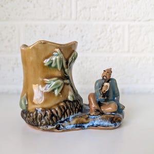 Puede incluir: Una maceta de cerámica con forma de tronco de árbol con una pequeña figura de un hombre sentado en la base. La maceta es marrón con hojas verdes y la figura lleva ropa azul.