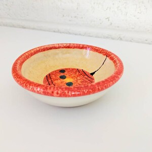 May include: A small, white ceramic bowl with a red rim and a hand-painted design of a red and yellow circle with black dots inside. The bowl is perfect for serving small snacks or dips.