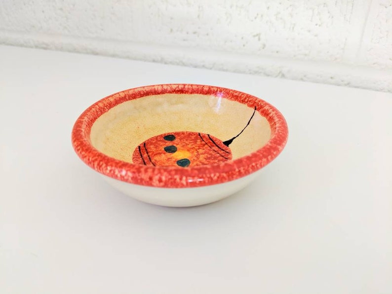 May include: A small, white ceramic bowl with a red rim and a hand-painted design of a red and yellow circle with black dots inside. The bowl is perfect for serving small snacks or dips.