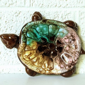 May include: A ceramic turtle dish with a brown, green, and pink glaze. The dish is shaped like a turtle with a raised shell and four legs.