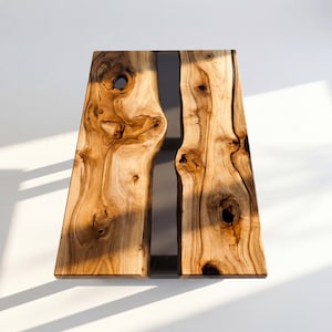 Op de afbeelding: Een handgemaakte houten tafel met een natuurlijke houtnerf afwerking. De tafel heeft een donkere, rechthoekige harsinleg in het midden. Het hout heeft knoesten en unieke patronen, wat een rustieke en moderne uitstraling creëert.