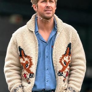 May include: A cream-colored, knitted cardigan sweater with a shawl collar. The sweater features a fox design in brown, black, and red on the front. The person is wearing a blue button-down shirt and blue jeans.