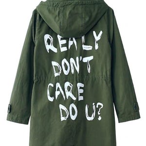 Puede incluir: Una parka con capucha de color verde oliva con mangas largas con puños abotonados y cintura con cordón. La parte posterior de la chaqueta muestra el texto pintado en blanco "REALY DON'T CARE DO U?" en una fuente de estilo manuscrito, audaz y desgastada.