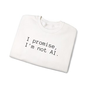 Op de afbeelding: Een witte sweatshirt met de zwarte tekst "I promise, I'm not AI." Het kledingstuk is opgevouwen, wat erop wijst dat het nieuw of vers gewassen is. Het ontwerp is minimalistisch, waardoor het een veelzijdig kledingstuk is voor dagelijks gebruik.