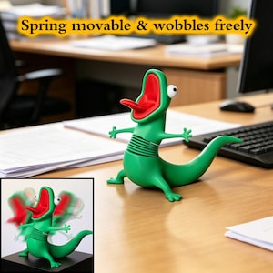 Puede incluir: Un juguete de escritorio con forma de lagarto verde con boca y lengua rojas, y ojos blancos. El juguete tiene un muelle y está sobre un escritorio de madera. El texto "Spring movable & wobbles freely" está encima.