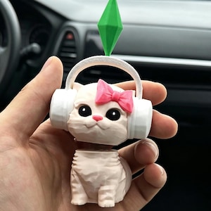 May include: A light pink cat figurine with a pink bow, white headphones, and a green diamond-shaped charm. The cat has large black eyes and a small pink nose. The figurine is designed to bobble.