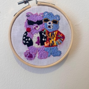 May include: Embroidered hoop art featuring two cartoon bears wearing sunglasses and stylish outfits. One bear is purple with a black jacket and the number 4. The other is blue with a colorful sweater. The artwork is mounted in a wooden embroidery hoop.