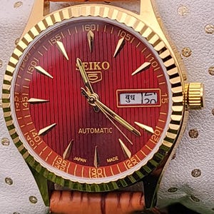 Peut inclure: Montre automatique Seiko 5 dorée avec un cadran rouge et un bracelet en cuir marron. Le cadran affiche le jour et la date, avec des aiguilles et des marqueurs dorés. La montre mesure environ 3,5 cm de diamètre.