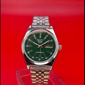 May include: A silver-toned Seiko automatic wristwatch with a green face and a silver band. The watch face displays the day and date, with the word "Automatic" printed below the center. The watch is set against a red background.