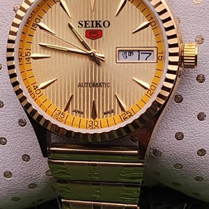 May include: Gold-toned Seiko automatic wristwatch with a textured dial and a date display. The watch features a gold-colored band and a fluted bezel. The watch is displayed on a white surface with a green background.
