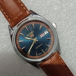 May include: A Seiko 5 automatic wristwatch with a round silver-tone stainless steel case and a textured blue dial. The dial features gold-tone hands, hour markers, a day-date window displaying SUN 7, and a brown leather strap with a crocodile pattern.