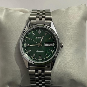 Peut inclure: Une montre-bracelet automatique Seiko en acier inoxydable avec un cadran vert. La montre affiche le jour et la date à 3 heures et possède un bracelet argenté. La montre est présentée sur un coussin blanc.