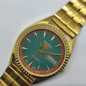May include: Gold-toned Seiko automatic watch with a teal dial and gold hands. The watch face features red and gold accents, with the day displayed. The watch band is also gold-toned.