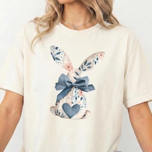 以下が含まれることがあります： クリーム色のTシャツ。花柄のウサギのデザイン。ウサギは模様入りの耳、青いリボン、ハート型のディテールがあります。デザインにはピンクと青の花柄の要素が組み込まれており、柔らかく春らしい美しさを生み出しています。