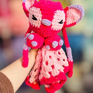 May include: A handmade, plush toy in shades of pink and lavender. The crochet toy features large ears, closed eyes, and a patterned face. The toy has a blanket-like bottom with a scalloped edge and is held by a person.