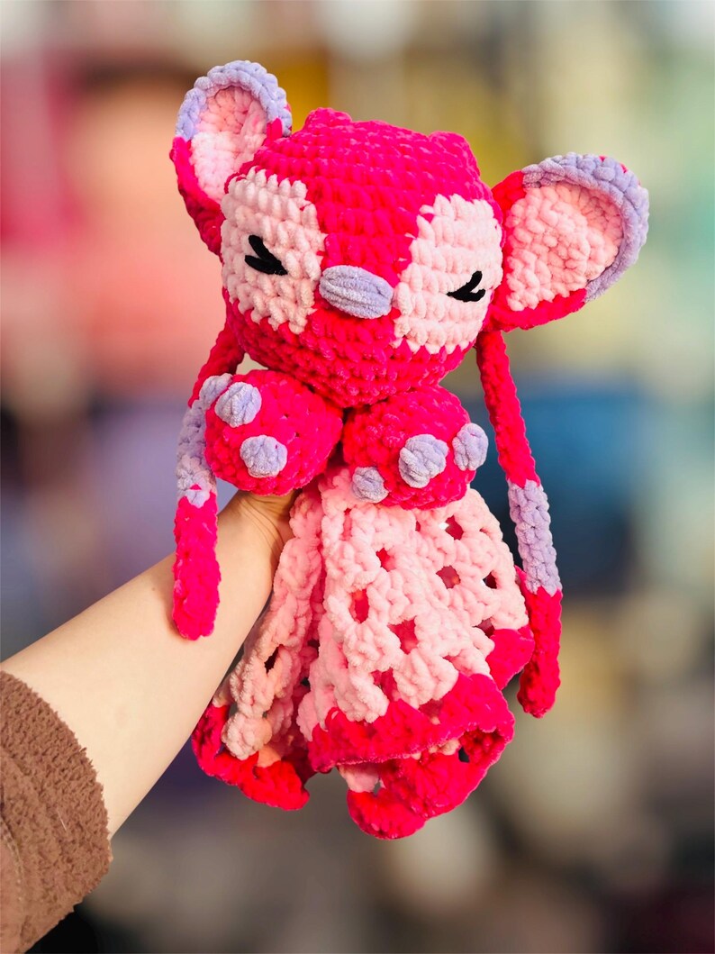 May include: A handmade, plush toy in shades of pink and lavender. The crochet toy features large ears, closed eyes, and a patterned face. The toy has a blanket-like bottom with a scalloped edge and is held by a person.