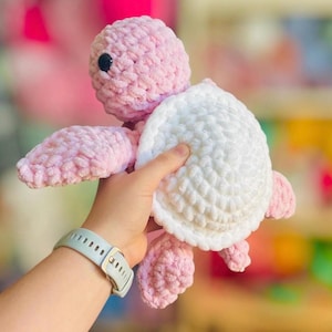 May include: A handmade crochet turtle plushie in soft pink and white. The turtle has a pink head, flippers, and feet, with a white shell. The plushie is held in a person's hand, showcasing the texture and detail of the yarn.
