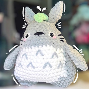 May include: A gray and white crocheted plush toy featuring a rounded body, pointed ears, and a light green tuft on top. The creature has black eyes with white highlights, pink cheeks, and a white belly with gray chevron-shaped markings.