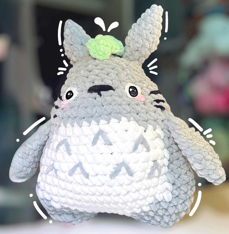 May include: A gray and white crocheted plush toy featuring a rounded body, pointed ears, and a light green tuft on top. The creature has black eyes with white highlights, pink cheeks, and a white belly with gray chevron-shaped markings.