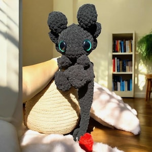 May include: A black crocheted dragon plushie with large, round, teal-rimmed eyes and a red heart-shaped tail. The dragon has large, round ears and is made of a soft, textured yarn. The plushie is sitting on a beige cushion.