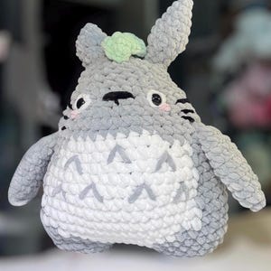 May include: A plush toy crafted from grey and white yarn, featuring a rounded body, pointed ears, and a light green accent on the head. The white belly displays grey chevron patterns, while the face has black eyes, whiskers, and pink cheeks.