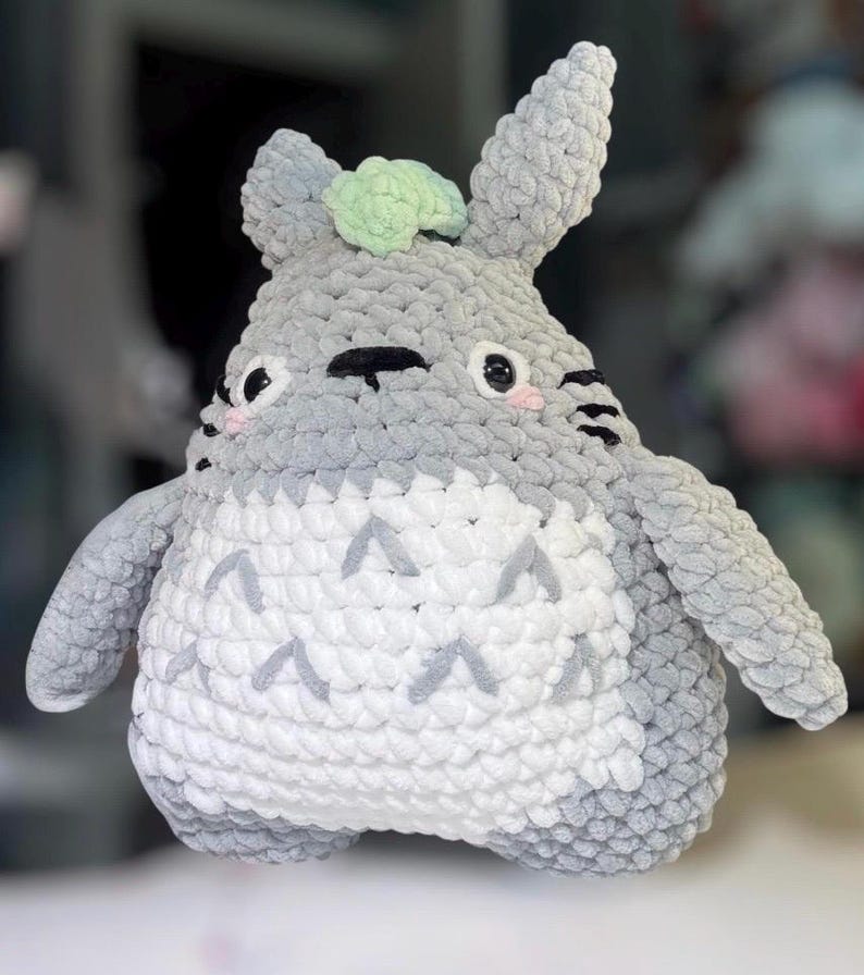May include: A plush toy crafted from grey and white yarn, featuring a rounded body, pointed ears, and a light green accent on the head. The white belly displays grey chevron patterns, while the face has black eyes, whiskers, and pink cheeks.