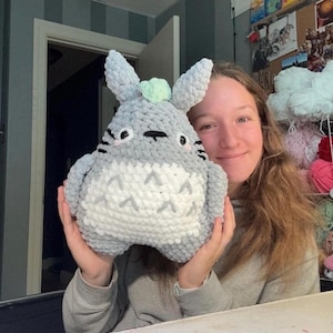 May include: A plush, crocheted stuffed animal featuring a rounded gray body with a white belly, pointed ears, and a small light green accent on top. The character has black embroidered eyes, whiskers, and a chevron pattern on its white chest.