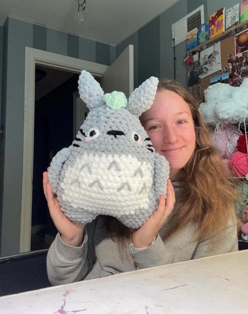 May include: A plush, crocheted stuffed animal featuring a rounded gray body with a white belly, pointed ears, and a small light green accent on top. The character has black embroidered eyes, whiskers, and a chevron pattern on its white chest.