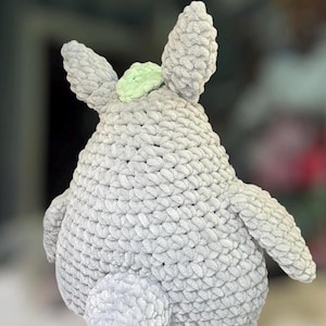 May include: A plush toy crafted from thick, light gray yarn with a textured, looped stitch pattern. The figure features two pointed ears, a rounded body, two side arms, and a small, pale green accent piece positioned between the ears.