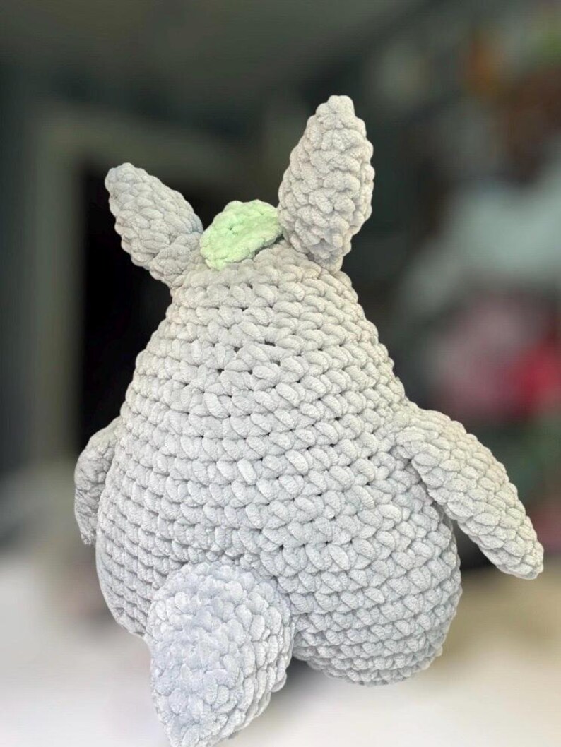 May include: A plush toy crafted from thick, light gray yarn with a textured, looped stitch pattern. The figure features two pointed ears, a rounded body, two side arms, and a small, pale green accent piece positioned between the ears.