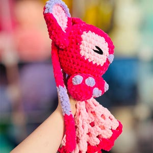 May include: A handmade, crochet bunny in shades of pink, lavender, and white. The bunny features long ears, a bow with purple polka dots, and a decorative openwork skirt. The bunny is held up by a person's hand.